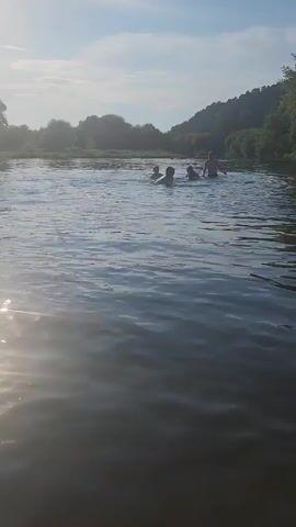 A group of people are swimming in a body of water. The water appears to be a river or lake, with trees and a hill in the background. The sun is shining, casting a bright light on the water. Some of the people are partially submerged in the water, while others are swimming. The scene is peaceful and relaxing.