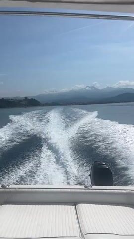 The video captures a boat ride on a bright, sunny day off the coast of Marina di Casal Velino, Italy. The primary view is from the stern of a white powerboat, showing its foamy white wake stretching behind it on the clear blue water. In the distance, a coastline with green hills, scattered buildings, and a range of mountains partly covered by clouds are visible under a clear blue sky.

A man, seen from the chest up, is onboard the boat, wearing a green t-shirt and a necklace. He holds a dark smartphone in his hands, with the sparkling sea and coastline in the background, appearing relaxed. Later, a close-up shows the man's hand gesturing, revealing light-colored, patterned shorts. The video then returns to a shot of the boat's wake and the receding coastal scenery, as the man's hand gestures towards the view. An outboard motor is visible at the stern of the boat, which features white padded seating. The overall impression is one of a leisurely and enjoyable trip on the water. No visible text is present in the video.