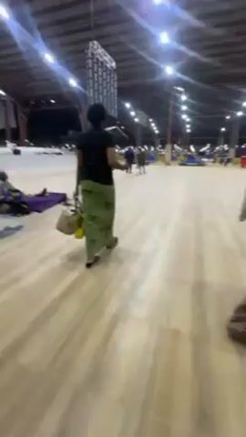 The video shows a large indoor space, possibly a warehouse or a large community hall, with a high ceiling supported by metal beams. The floor is covered with light-colored, possibly wooden, planks. The space is well-lit by rows of overhead fluorescent lights, creating a bright environment.

Several people are present in the video. In the foreground, a child is sitting or lying on a blanket, with what appears to be a pillow or bag nearby. Further ahead, a person wearing a dark shirt and green pants is walking away from the camera, carrying a bag or purse. As the camera moves forward, two women dressed in traditional African clothing come into view, walking in the same direction. The woman in the middle is wearing a patterned grey and brown garment, while the woman on the right is dressed in yellow and purple attire.

In the background, there are more people scattered throughout the space, some appearing to be sitting or lying down, while others are walking around. The overall atmosphere suggests a temporary or communal gathering, possibly related to a religious or community event. The space seems to serve as a sleeping or resting area for the attendees.
