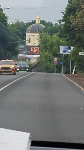 This is a first-person perspective video of a vehicle driving on a paved road. The road has a double yellow line in the center. On the left side of the road, there is a billboard with an advertisement. Further down the road on the left, there is a green vehicle. On the right side of the road, there are multiple road signs, including directional signs and regulatory signs. Beyond the road, there are trees and a building with a dome. The lighting suggests it is daytime. There are no people clearly visible.