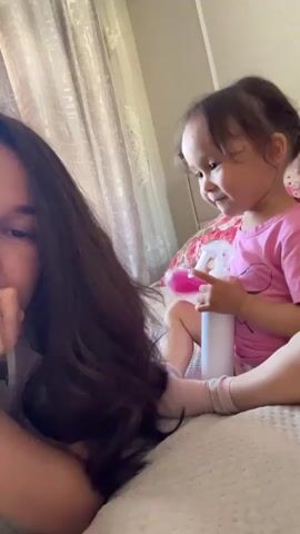 A video shows a young girl and a woman on a bed. The woman is lying down facing the camera, with her head propped up by her hand. She has long, dark brown hair. Next to her, a young girl, dressed in a pink t-shirt, sits cross-legged, holding a white spray bottle. She smiles and laughs, seemingly playing with the bottle. She has her hair in two small braids. The background includes a bed with a patterned headboard and curtains hanging behind it. The video is taken from a close-up perspective, focusing on the faces of the woman and the child.