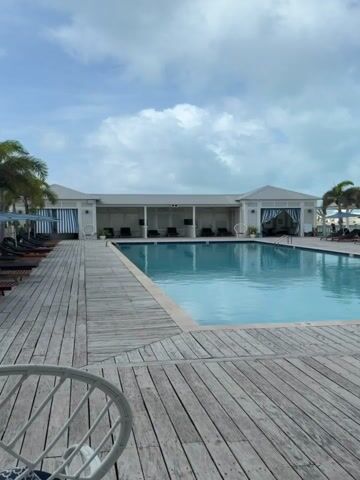 The video presents a static, wide shot of an empty outdoor swimming pool area. A large rectangular swimming pool with clear blue water occupies the center. The pool is bordered by a light-colored wooden deck. On the far side of the pool, a long, white, single-story building with a pitched roof and multiple open cabana-like sections is visible. These sections contain dark lounge chairs. The building has a central pavilion area with blue and white striped curtains. To the right of the pool, several dark lounge chairs are arranged, some with umbrellas, and a few palm trees are scattered. The sky above is partly cloudy with visible blue patches. In the bottom left foreground, a portion of a white wicker chair is visible, and on the bottom right, a part of a brown and white striped cushion or seat is seen. No people are present in the scene.
