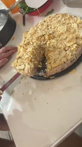 A close-up shot shows a hand using a knife to cut a slice from a large, layered cake. The cake is covered in flaky, golden-brown pieces, likely from crushed pastries or cookies, and appears to have a creamy filling with visible hints of red fruit. The cake is on a black serving plate, which sits on a white tablecloth. In the background, out of focus, are various objects on a table including an orange plastic cup, a red and white bowl with green leafy vegetables, a white lid, and part of a silver baking pan. The lighting is dim, suggesting an indoor setting.