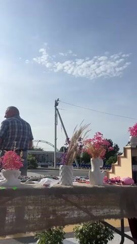 A man in a plaid shirt and jeans walks past a stall displaying various items, including dried flowers in vases. In the background, a stage is set up with a band performing and a Ferris wheel can be seen. The sky is clear and blue. The man is holding a pink shopping bag.