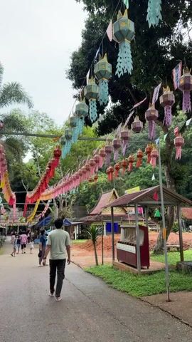 A wide outdoor shot shows a narrow paved road or path with people walking on it. The path is decorated with colorful lanterns and banners hanging overhead. The lanterns are of various shapes and sizes, mostly red, pink, and light blue. The path is lined with trees and small buildings on both sides. A vendor is speaking in the background.