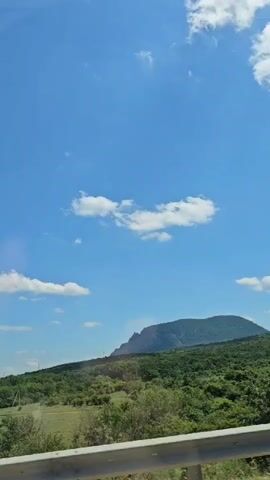 This media file shows a scenic view from a moving vehicle, likely a car, along a road in Krasny Pakhar, Russia. The primary subject is a large, verdant mountain range under a bright blue sky dotted with fluffy white clouds. The foreground features a silver metal guardrail, suggesting the viewer is on a highway or a similar roadway.

The scene is bathed in daylight, indicating it is daytime, likely late morning or afternoon given the brightness and the presence of shadows. The weather appears to be clear and pleasant, with the sun shining and casting light on the landscape. The mountain is covered in dense green foliage, with trees and shrubs creating a rich texture. The lower parts of the landscape include grassy fields and scattered trees.

There are no people or specific objects visible beyond the natural scenery and the guardrail. The activity taking place is implied: travel. The viewer is observing the passing landscape from inside a vehicle. The overall emotion conveyed is one of peacefulness and appreciation for nature's beauty. There is no visible text within the media file. The location context of Krasny Pakhar, Russia, places this natural vista in a specific geographical setting.