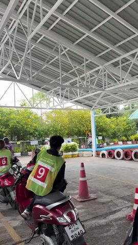 Several people wearing numbered vests are waiting on scooters under a metal-roofed structure. Outside the structure, there are trees, and an area demarcated by traffic cones and stacks of tires. A sign in Thai advertises car tax and insurance consultation. One person is talking on their phone while sitting on a scooter. License plates are visible on the scooters.