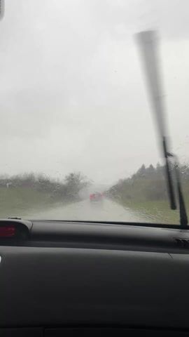 The video shows a car driving on a wet road. Rain is visible on the windshield, and the windshield wipers are in motion. The car is following another vehicle, visible in the distance. The dashboard of the car is visible, including the speedometer, radio, and air conditioning vents. There is an air freshener hanging from the rearview mirror. The car appears to be driving on a rural road with trees on either side. The sky is overcast and grey.