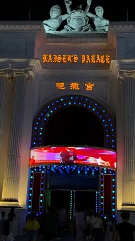 The video shows the entrance of a building with a sign that reads "KAISER'S PALACE". Above the entrance, there are statues and architectural details. The entrance itself is an arched doorway decorated with a marquee of red and blue lights. Below the marquee, a large LED screen displays colorful images, featuring people in various poses and outfits. A dark red curtain is visible behind the screen, and silhouetted figures of dancers are projected onto it. A crowd of people, dressed in casual clothing, are walking towards the entrance. The scene appears to be at night, with artificial lighting illuminating the building and the surrounding area. The overall impression is that of an entertainment venue.