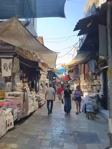 The video shows a busy outdoor market. People are walking down a cobblestone path lined with shops on both sides. The shops are selling various goods, including textiles, clothing, and souvenirs. Sunlight filters through awnings and canopies, creating a dappled effect. The atmosphere appears lively and bustling.