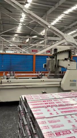 The video shows a machine cutting a metal bar in an industrial setting. The machine is large and white, with a cutting head that moves along a track. The bar is long and silver and is secured in place. In front of the machine, there is a stack of packaged metal plates. The setting is a large factory with exposed metal beams and bright lights. The walls are partially covered with blue and orange safety barriers. The machine operates autonomously, performing repetitive cutting actions.