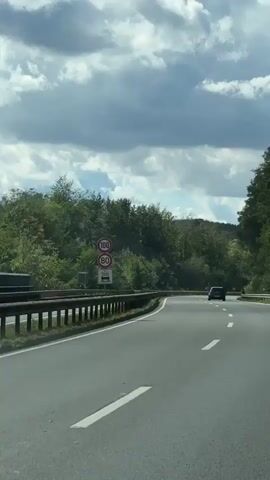 A dark-colored car drives on a paved road with a white line marking the lane. The road curves to the right, flanked by a metal guardrail on the left and trees on both sides. Ahead, there are speed limit signs indicating 100 and 80. The sky is partly cloudy.
