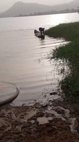 The video shows a fisherman pushing a small canoe into a calm lake on an overcast day. A pump is sitting on the muddy bank of the lake, a gray hose leading into the water. A small hill is visible in the background.
