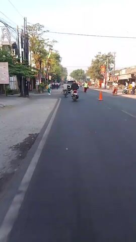 A street view, taken from a moving vehicle, shows a road with a sidewalk on the left and a group of people walking on the sidewalk. Several motorcycles are also visible on the road, moving in the same direction as the camera. Trees line the street, and buildings are visible on the left side. The sky is clear and bright, indicating daytime. Traffic cones are placed on the right side of the road.