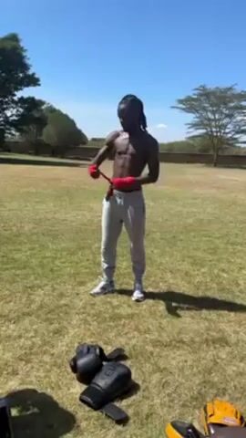 A slender, dark-skinned man with dreadlocks, bare-chested and wearing grey sweatpants and athletic shoes, is wrapping his hands in red boxing wraps. He is standing in a grassy field on a sunny day. Some boxing gloves are sitting on the ground near him. In the background are some trees and a stone fence.