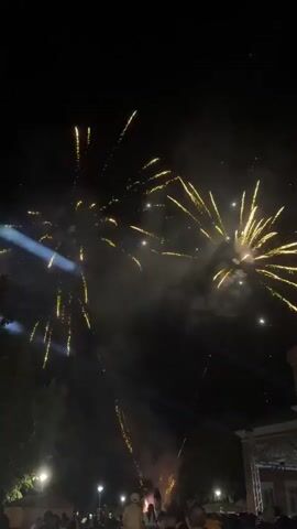 The video shows a nighttime scene of a fireworks display. Several bursts of yellow and white fireworks are visible against a dark sky. Smoke from the fireworks drifts across the sky. In the foreground, silhouettes of people are visible, gathered to watch the display. Some trees are also visible on the left side of the frame. The bottom right corner shows a building with some lights. The video captures the festive atmosphere of the event.