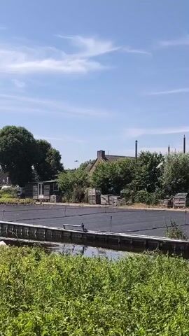 A video shows a bird in flight against a blue sky with scattered clouds. In the foreground, lush green vegetation grows along a body of water. A wooden pier with a bench crosses the water, and a dark, flat surface, possibly a tarp or covered area, is visible behind it. Beyond the pier, a small building with a gabled roof and a chimney is surrounded by trees and wooden crates. The overall scene depicts a rural or semi-rural outdoor environment on a sunny day.
