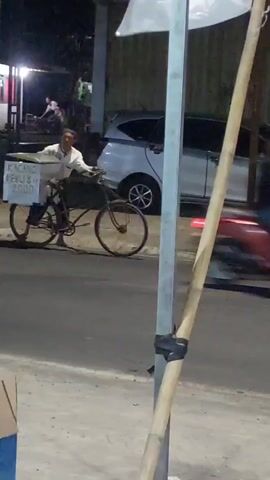 A man is riding a bicycle at night. He is wearing a light-colored shirt and a hat. A box is attached to the front of his bicycle, and it has writing on it. The writing on the box says "KACANG REBUS" and "RP 2000". The man is riding on a paved road, and there is a white car behind him. There are also streetlights and buildings in the background. The video appears to be taken from a stationary position.