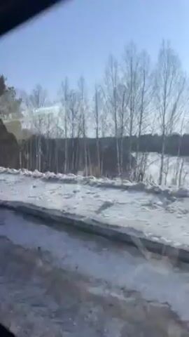 The video shows a view from a moving vehicle, likely a car or bus, traveling along a snow-covered road. The landscape outside the window features trees, some bare and some with foliage, and snow-covered ground. The sky is bright and clear. The window frame is visible at the edges of the frame. The camera angle remains steady, focused on the passing scenery.
