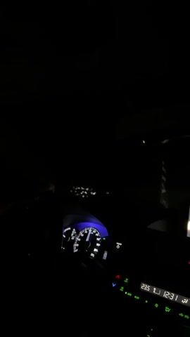 The video shows the interior of a car at night. The dashboard lights up the driver's side, displaying a speedometer and other indicators. To the right, a screen shows song information and track duration. Outside the car, distant lights suggest a road or urban environment. The overall impression is of a nighttime drive.