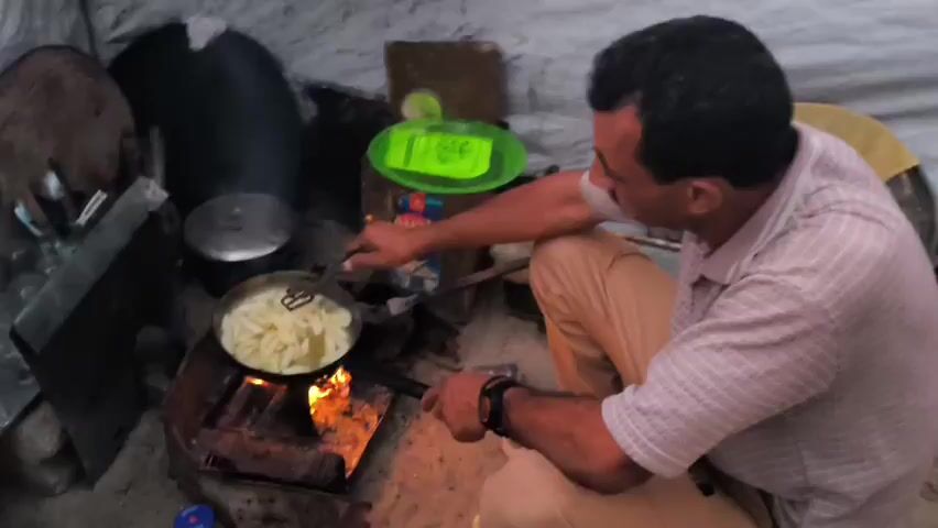 The video shows a person cooking french fries in a pan over an open fire. The person is holding two tongs and stirring the fries in the pan. The fire is burning brightly beneath the pan, and there is smoke rising from the fire. The background is blurry, but it appears to be an outdoor setting.