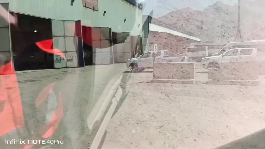 The video opens with a blurry, reflected view of a scene, possibly through a window or windshield. We see concrete barriers, a white truck, and buildings in the background, all set against a backdrop of mountains. The timestamp reads 00:00. The scene then shifts to a clearer, wider shot. A clear sky stretches above rugged, layered mountains. In the foreground, there are several small buildings with red-tiled roofs. To the left, a large white structure, possibly a warehouse or factory, is visible. A yellow gantry crane stands near the white building. Further to the right, a yellow excavator is parked. The timestamp is now 00:01 and progresses to 00:05, showing the same scene. There are no people visible in the video. The environment appears to be an industrial or construction site located in a mountainous, arid region.