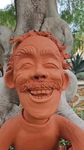 The video shows a close-up of a terracotta sculpture of a grinning man with a mustache, sitting in a lush green garden. The sculpture is placed in front of a large tree with sculpted grey elements that resemble elephant legs. In the background, a dinosaur sculpture and other plants are visible. The camera slowly zooms out, revealing more of the garden environment and the scale of the sculptures.