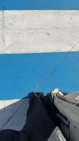 From a first-person perspective, a person walks on a cracked concrete surface with alternating painted stripes of blue and white. The person is wearing black pants and pointed black shoes. A rolling suitcase with a white body and black wheels is pulled along beside them. The environment appears to be outdoors in bright sunlight. The video is handheld and the movement is natural, following the walking motion.