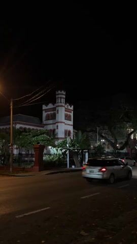 At night, a low-angle, static shot shows a roadway with a building on the other side. The building has a tall tower structure painted white and trimmed with reddish-brown. The main building structure is also painted in the same colors, with the trim around the windows highlighted. The building is surrounded by a black fence and some trees. On the roadway, two white vehicles drive by at different times. A lamp post is briefly centered in the frame. The sky is completely black. The scene is dark and the street lighting is minimal.