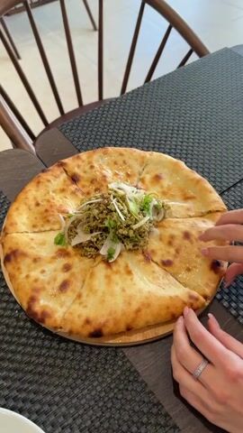 A close-up shot shows a round, golden-brown flatbread topped with a mixture of green ingredients, onions, and cilantro. The flatbread is cut into eight equal slices, and one slice is being lifted by two hands. One hand, with a ring on the ring finger, is holding the crust of the slice, while the other hand, with a diamond ring on the ring finger, is supporting the underside of the slice. The flatbread is on a wooden board, which is placed on a dark gray placemat. In the background, the legs of a wooden chair are visible. The lighting is bright, suggesting an indoor setting.