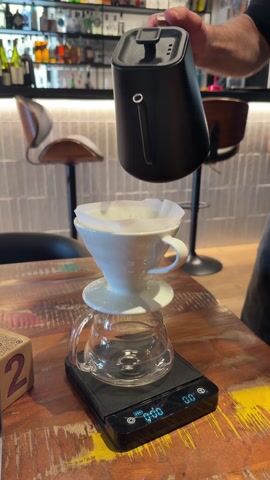 A man is pouring water from a black kettle into a white ceramic pour-over coffee maker set on top of a clear glass pitcher, all placed on a black digital scale. The scale sits on a distressed wooden table with colorful paint.  Behind the setup are two bar stools with leather seats and metal bases, and a bar area with liquor bottles on display.  A wooden cube with the number "2" is visible on the left side of the frame.  The man’s hand and arm are visible as he pours the water with a slow, steady stream. The scale is displaying the weight in grams of the water being poured.