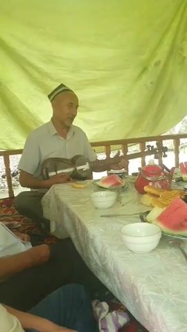 A man wearing a patterned cap sits cross-legged on the floor, playing a stringed instrument. He is seated at a table laden with food and drink. Slices of watermelon, bowls of white liquid, and a red teapot are visible. The man is dressed in a light-colored collared shirt and dark pants. He appears to be singing or speaking while playing, his mouth open and eyes looking forward. He is positioned under a pale green canopy, with wooden railings visible in the background. The overall setting suggests an outdoor gathering, possibly a meal or celebration. The lighting indicates it is daytime. The man's expression seems engaged and perhaps joyful. There is no visible text in the image.