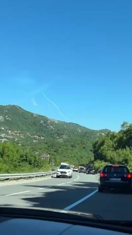 The video shows a view from the inside of a car driving on a road. The road has multiple lanes and is bordered by green mountains and trees. Several cars and a white van are visible on the road ahead. The sky is clear and blue with a few wispy clouds. The time stamp indicates the video is from January 1, 2023.