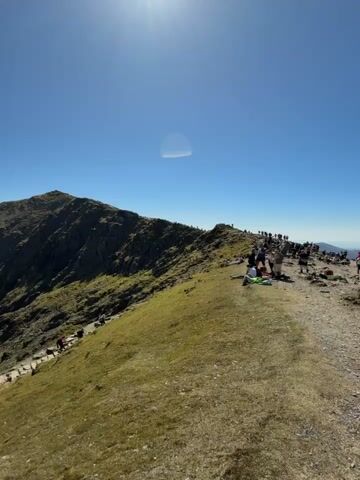 The video shows a mountain landscape under a clear blue sky with a bright sun. People are sitting and standing on the mountain's peak. A path leads down the mountainside, with people walking along it. In the distance, two lakes are visible in the valley.