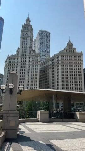 A video pans from right to left showing a cityscape on a clear day. In the foreground on the left, a concrete pillar with three lights is visible. Behind it is a contemporary glass-enclosed structure with a curved roof. The middle ground features a large, ornate white building with a clock tower and a connected, longer white building. To the far left, a modern blue glass skyscraper is visible. The sky is a bright, clear blue.