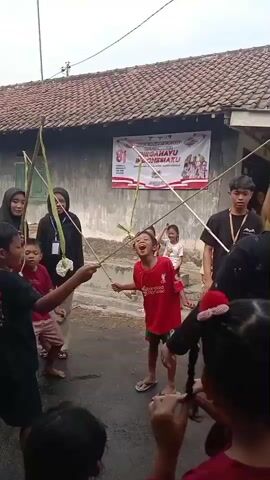 A group of children and adults are gathered outdoors, possibly in a street or yard, for what appears to be a game or event. In the center, a young boy in a red t-shirt and green shorts is participating in a challenge. He is standing with his mouth open, attempting to eat something suspended from a long, thin stick held by two people. The stick is attached to a larger, X-shaped structure made of bamboo or similar material, held up by multiple people. Several other children are watching, some actively participating in holding the structure. In the background, a banner with Indonesian text, possibly celebrating an anniversary ("DIRGAHAYU INDONESIA"), is displayed on a building. The environment appears to be somewhat overcast. The overall atmosphere seems festive and communal, likely related to a local celebration or holiday.