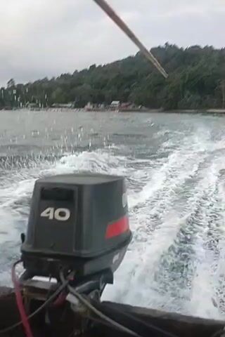 The video shows the stern of a boat moving through choppy water. An outboard motor with "40" printed on its casing is visible. A wooden pole extends upwards from the boat and out of frame. In the distance, a treed hillside with some buildings is visible along the coastline. The sky is overcast.