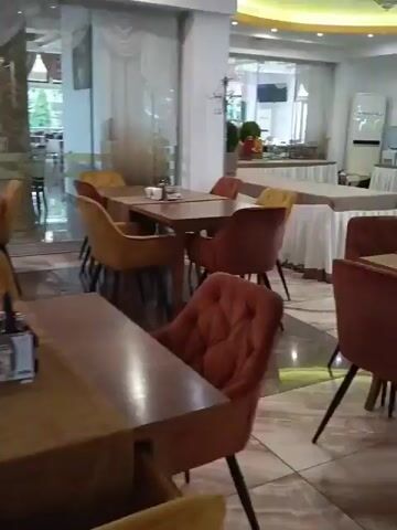 The video shows the interior of a restaurant or cafe. There are many tables and chairs, most of which are empty. The tables are set with tablecloths, placemats, and some have salt and pepper shakers. The chairs are upholstered in warm colors like orange and yellow. The room appears to be well-lit with artificial lighting. In the background, there are large windows with a view of what appears to be an outdoor area with greenery. There are also some service counters and possibly kitchen equipment visible in the background. The overall atmosphere suggests a modern and somewhat elegant establishment.