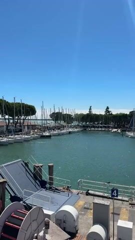 The video shows a marina on a clear, sunny day. In the foreground, there is a ramp with a dark surface leading down to the water. To the left of the ramp, there is a large spool with red and black accents. Several wooden posts are visible around the ramp. The water is a vibrant turquoise color and appears calm. In the background, a row of sailboats with tall masts are docked. Behind the sailboats, there are buildings with red roofs and a line of trees with distinctive, umbrella-shaped canopies. The sky is a bright, clear blue.