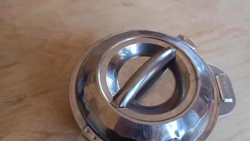 A person removes the lid of a stainless steel pot, revealing a small piece of dark brown, possibly burnt, food residue inside. The pot has a slightly tarnished appearance, indicating use. The background is a light-colored wooden surface.