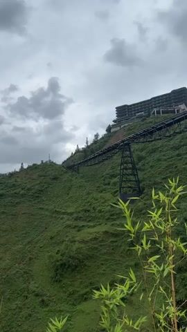 The video depicts a lush green hillside under a cloudy sky.  A black metal bridge-like structure supported by vertical supports runs across the slope. A large, multi-story building sits at the top of the hill, connected to the bridge structure. In the foreground are some out-of-focus green plants.