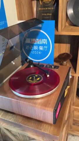A close-up shot of a turntable playing a red vinyl record. The turntable has a wooden base and a silver tonearm. Behind the turntable are two blue circular signs with Chinese characters and the text "2024". To the right of the turntable is a wooden shelf filled with awards and vinyl records. The lighting is warm and indoor.