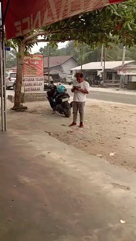 The video shows an outdoor scene in what appears to be a commercial or roadside area. A man stands near a motorcycle, looking at his phone. He is wearing a white t-shirt, gray pants, and red shoes. To the left, a woman walks past, carrying a bag over her shoulder. She is wearing a patterned shirt and jeans. In the background, there are buildings, other vehicles, and trees. A red banner hangs overhead with white lettering. There is a sign board displaying text in Indonesian, mentioning food items like "Martabak Telor" and "Roti Bakar." The ground is a mix of concrete and dirt.