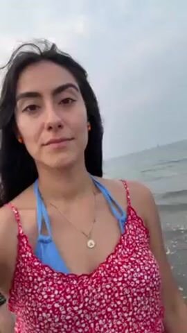 A young woman is recording a selfie video on a beach. She is wearing a red and white floral patterned dress over a blue bikini top. She has long dark hair that is blowing in the wind.  She is wearing a necklace and a bracelet. The ocean is behind her, with waves visible. The sky is overcast. She smiles at the camera and runs her hand through her hair.