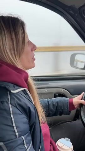 The video shows a woman driving a car on a foggy day. The woman has long blonde hair and is wearing a dark jacket and a maroon hoodie. She has a small cup with a blue lid on the passenger seat. The car has a black steering wheel and a phone in the dashboard displaying a map. The windshield wiper is visible, and a purple hat sits on the dashboard. The road has a yellow guardrail. The weather outside is foggy and the visibility is low.