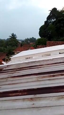 The video shows an aerial view of a residential area. The foreground is dominated by the corrugated metal roof of a building. Beyond this, there are several houses with red tile roofs, interspersed with trees and foliage. In the background, more houses and trees are visible, with a hazy sky overhead. The scene appears to be a somewhat rural or semi-rural environment. The camera is panning slowly from left to right, capturing the expanse of the neighborhood. There are no people or animals visible in the frame. The lighting suggests it might be overcast or late afternoon.