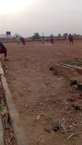 The image depicts an outdoor soccer game in progress on an unpaved, dusty field. The ground is a reddish-brown earth, appearing dry and uneven. In the foreground on the left, a concrete drainage channel or curb is visible, filled with dry leaves, plastic waste, and other debris.

The main subjects are the individuals playing soccer. At least eight players are visible on the field, with six actively engaged in the central action. Players are dressed in bright sportswear, predominantly green and purple/dark red shirts. One player in a bright green shirt is dribbling a white soccer ball towards the right side of the frame, with other players positioned around them, indicating an ongoing match. On the far left, partially visible, is a person seated on the ground, likely observing the game.

In the background, several simple, single-story buildings with light-colored walls and dark roofs are scattered across the landscape. A water tank on a raised stand is visible above one of these structures. Sparse trees line the distant horizon. The sky is a pale, diffused light blue-white, suggesting either a late afternoon or early evening setting, given the soft illumination and lack of harsh shadows.

The scene conveys a casual, recreational atmosphere in what appears to be a rural or semi-urban community setting, consistent with Baganakwo, Nigeria. The activity is clearly a game of football, played with enthusiasm despite the rudimentary field conditions. There is no visible text in the image.