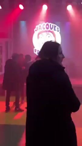 The video shows a group of people standing around in a dimly lit room. In the background, a screen displays an image of a person with the words "Jacques Fromage" written above their head. The image is surrounded by bright lights, and the room is filled with smoke.