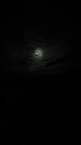 The video shows a dark night sky with clouds and the moon. The moon is partially obscured by clouds, giving it a hazy appearance. The lower part of the video is completely black, suggesting it is either a dark foreground or the video is focused on the sky. There are no people or identifiable objects visible in the frame. The overall impression is of a serene and slightly mysterious night scene.
