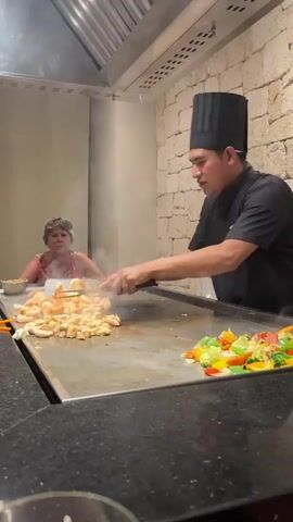 The video captures a chef in a black uniform and chef's hat cooking on a large flat-top grill in a restaurant. In the background, a woman wearing a floral-print dress is seated at the grill, observing the chef. The chef is actively cooking, stirring shrimp on the left side of the grill with tongs and preparing to add ingredients from a bowl to the right side, where mixed vegetables are already cooking. Steam rises from the grill, indicating the cooking process. The environment appears to be a teppanyaki-style restaurant with a stone-tiled wall in the background. The lighting suggests an indoor setting.